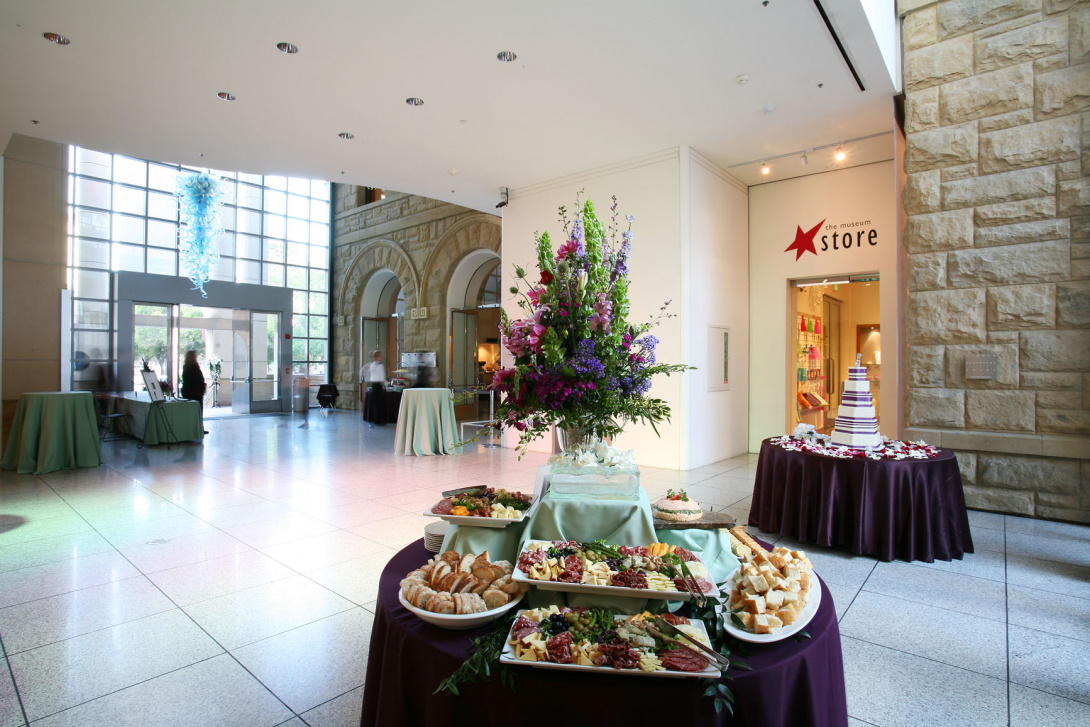 In a lobby with large stone walls, two tables with purple table clothes are stacked with food and flowers. Towards the back of the room, near the entrance are three tables with green table cloth.