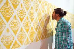 A male with his hair tied back stands in front of a wall covered with a yellow diamond pattern. Within each yellow diamond are 3 circles nested within each other, and a line that runs horizontal through the middle of them.