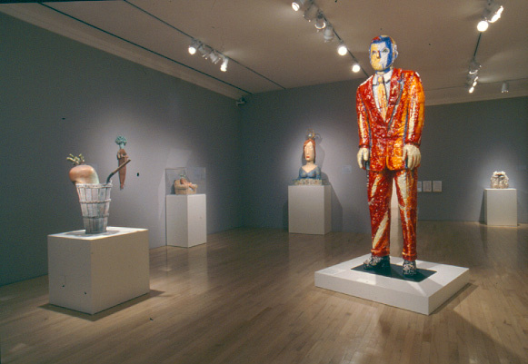 A gallery with grey walls, with multiple pieces on white pedestals near the walls, and one large sculpture of a man with an orange and red sequin suit. The man's face is bright blue and white to show shadows.
