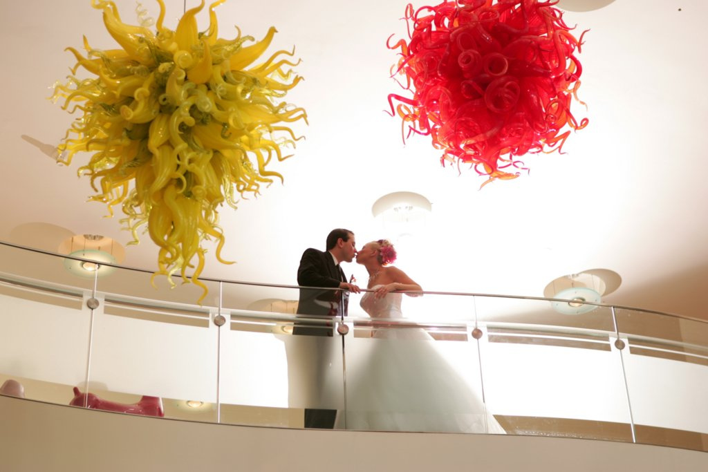 A man wearing black suit is kissing a female wearing a white wedding dress . She has a pink flower in hair. They are standing on a balcony overlooking one red and one yellow glass Chihuly chandelier.