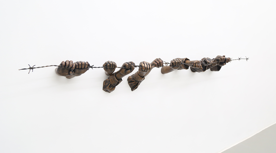 Bronze sculpure installed on a white wall. Sculputure is of hands holding on to barb wire.