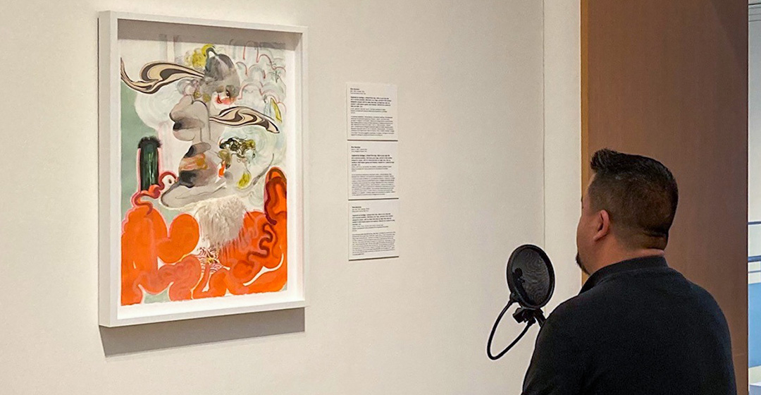 A person speaks into a microphone while viewing a framed artwork in a gallery. Wall text is displayed beside the piece.