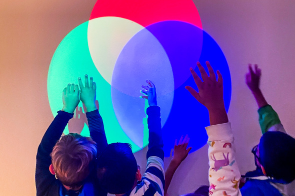 Three spotlights; red, green, and blue, shine on a wall demonstrating additive color mixing, the same technique used by computer screens. Children reach out their hands to block various colors, playing with the resulting colors.
