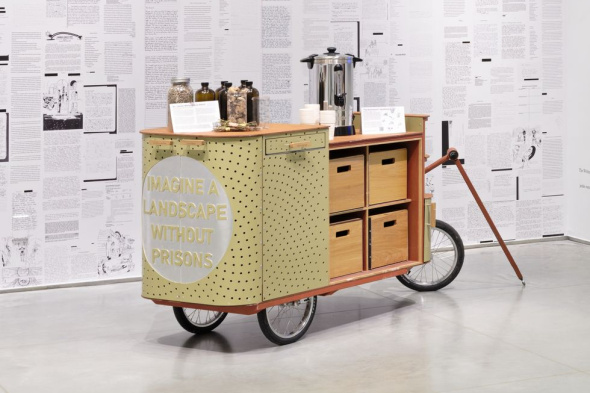 A yellow wooden mobile vendor cart with three wheels sits in the middle of a black and white room. The cart has a door open to reveal drawers, and jars of herbs and a hot water dispenser are placed on top. 