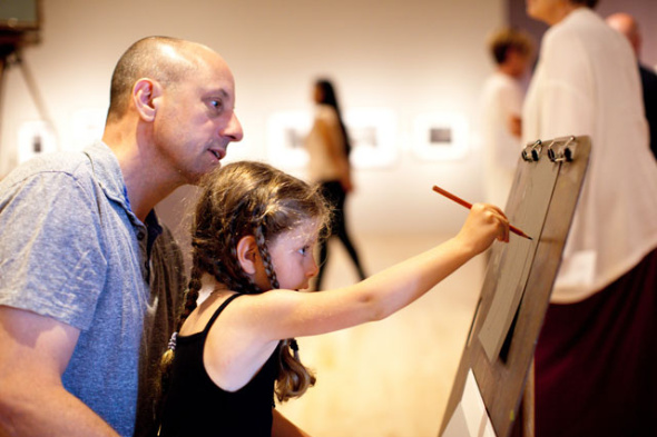 Sitting on her father’s lap, a young girl draws on an art easel in a gallery. People are in the background.