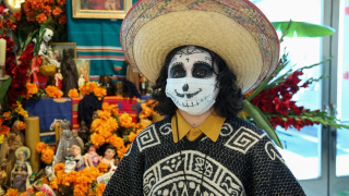 Person wearing skull makeup, a poncho, and a straw hat in front of an altar.