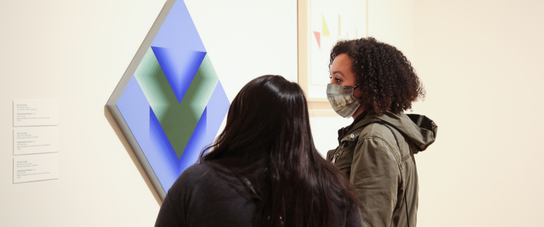 Two people look at an artwork. The artwork is square-shaped, with dark blue and grey "V"s against a powder blue background. The square painting is hung angled, with a point facing upwards, and another downwards. 