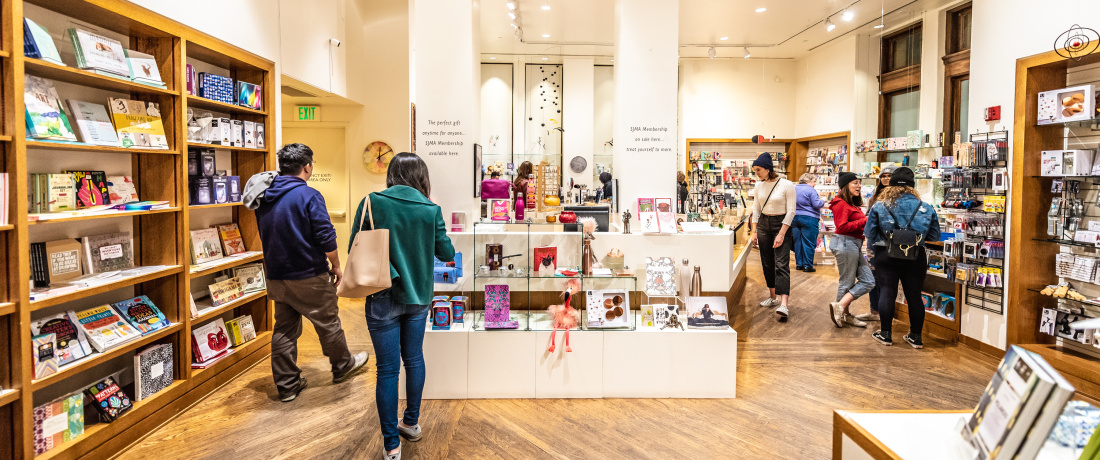 Wooden bookshelves line the walls of a store with nearly 10 people browsing at items or talking with one another. In the center of the room are white shelves with books and other objects. The floors are wooden.