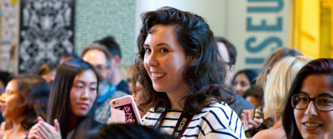 A young woman standing in a crowd with black wavy hair smiling,  Wearing a black and white striped shirt with a camera strap hanging around her neck and holding a phone with a pink phone case. People standing in the crowd are clapping