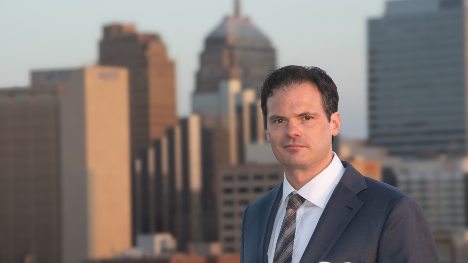 white man in a gray suit in front of a cityscape 