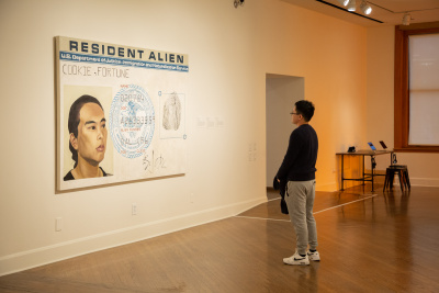 A person stands in a gallery viewing a large artwork resembling a Resident Alien card, featuring a portrait, fingerprint, and official seal