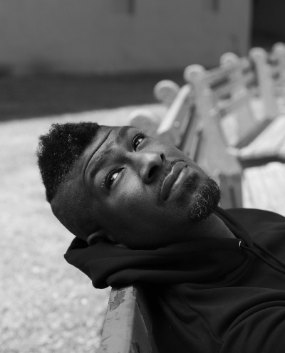 a photo portrait of a Black man with a faux hawk and chin beard lying back on a park bench gazing up