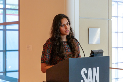 South Asian woman with long body waves with a floral patterned shirt speaking on a lectern