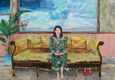 A barefoot woman wearing a long floral dress sits on a couch. Next to her is a large plant and behind her large windows show the blue sky. 