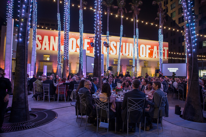 Dinner guests in formal attire are seated outdoors at round tables. Large palm trees are between the tables, each with small lights wrapped around the trunk. In the background, on the building, a large sign reads “San José Museum of Art.” 
