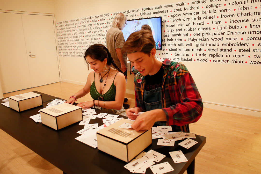 Two smiling young adults sit at a table covered in cards with words on them. The gallery wall has a screen affixed to it, along with a random list of words separated by bullet points. A man and another person look at the screen with—their backs to the viewer.