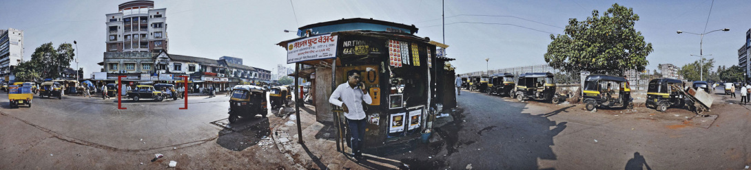 A photograph of a street view with a man on a phone in front of a small newsstand. He is in the middle of a long street lined with small cars. On the left side are several buildings and on the right side a fence lines the street with a tree at end of it.