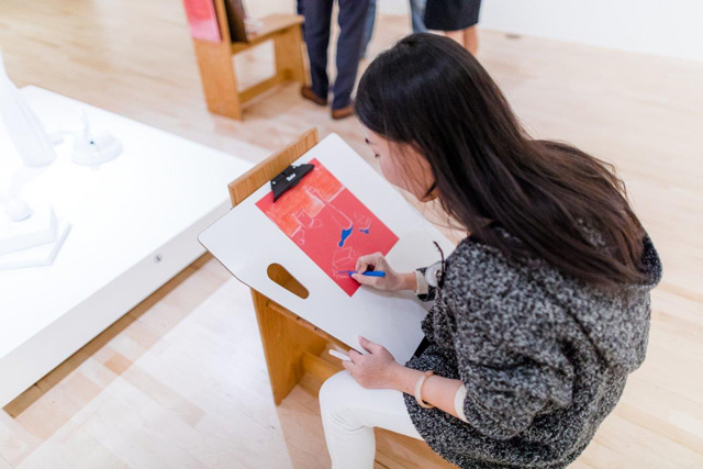 A young woman sits in a museum gallery. She looks at a sculpture while she draws in blue using an easel. Only the base of the sculpture on the pedestal is visible. Another chair is visible in the background and three sets of legs can be seen around it.