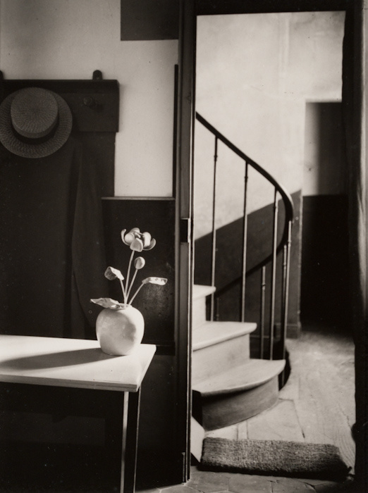 A black and white photograph of a doorway opens out to a hallway with a stairway that leads up and down. In front of the threshold is a ceramic potted flower. A brimmed straw hat hangs on the wall. Although there are several visible items, it feels empty.