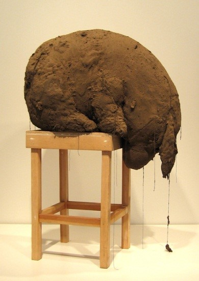 A giant piece of clay shaped like an animal that looks like Eeyore is sitting on a high wooden stool with four legs. The animal is looking down, leaning forward with its ears pointing downwards. Some of the dirt seems to be falling off it on the floor.