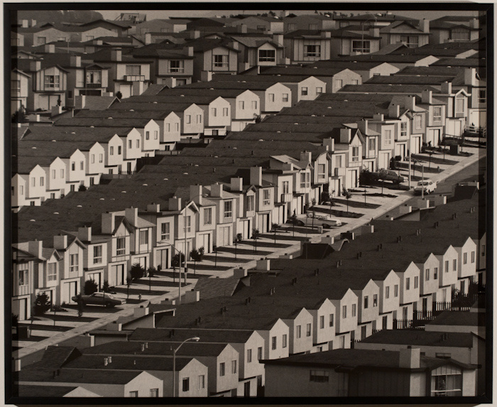 A sepia toned photograph of multiple rows of densely packed identical houses. They are lined up a hill with several cars from the late 1950s or 60s.