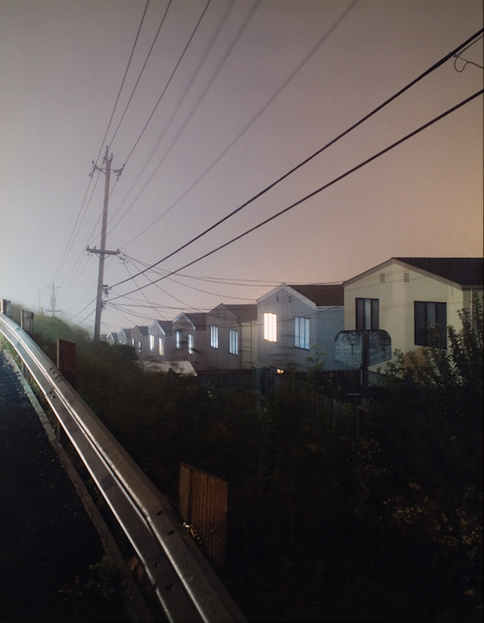 A photograph of a row of houses descend down a grade, behind a guardrail. Power lines parallel the houses with a faint glow of the sun behind the houses. 