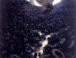 A photograph of a wasteland of rubber tires, they line to the top of each side of the photo. In the background is a flame with thick smoke coming out of it. The tires are on fire and the smoke pollutes the air.