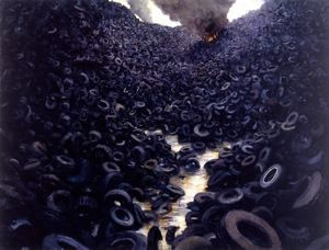 A photograph of a wasteland of rubber tires, they line to the top of each side of the photo. In the background is a flame with thick smoke coming out of it. The tires are on fire and the smoke pollutes the air.
