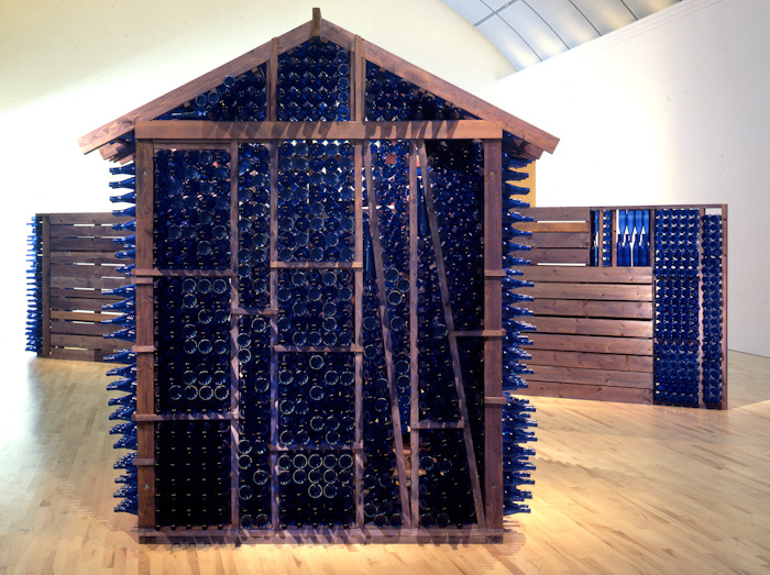 A wood framed house-like structure in front of wooden fences. The structure's walls are made with stacked blue glass bottles. The fences are made of wooden horizontal panels and on either ends of the walls are panels made from blue glass bottles.