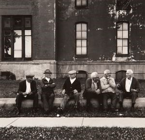 A black and white photograph of six elderly gentlemen siting in front of a building, talking to one other smiling. They seem to be having a good time. The building has 3 windows with a tree's reflection on the large window on the left side.