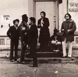 A black and white photograph of six young men of different ethnicity. They are dressed as though they are from the 1970s or 80s. They talk amongst each other. They stand except one who sits down on a stoop, smoking a cigarette and playing bongo drums.