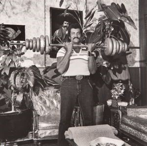 A black and white photo of a man in a tee shirt and jeans holds a weighted barbell at neck height in a nicely furnished living room. It appears to be taken in the 1970s.