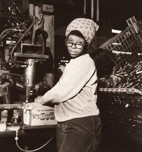 A black and white photograph of a black woman working in a factory setting. She stares at the camera.
