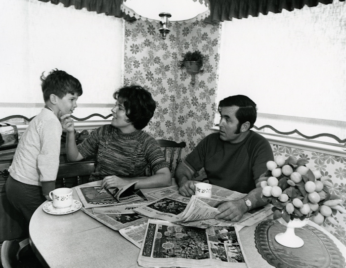 A black and white photo of a family around a kitchen table. The woman and child smile at one another while the woman holds the child's face in her right hand, as she flips through a paper. The man holds a newspaper and teacup while looking at them.