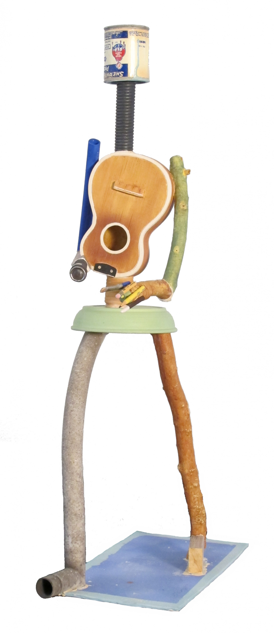 A porcelain sculpture consisting of various objects resembling a human. A can is the head, a black coiled tube is the neck, a guitar is the body, a bowl are hips, and the legs and arms are represented through a piece of wood and skinny metal tube.
