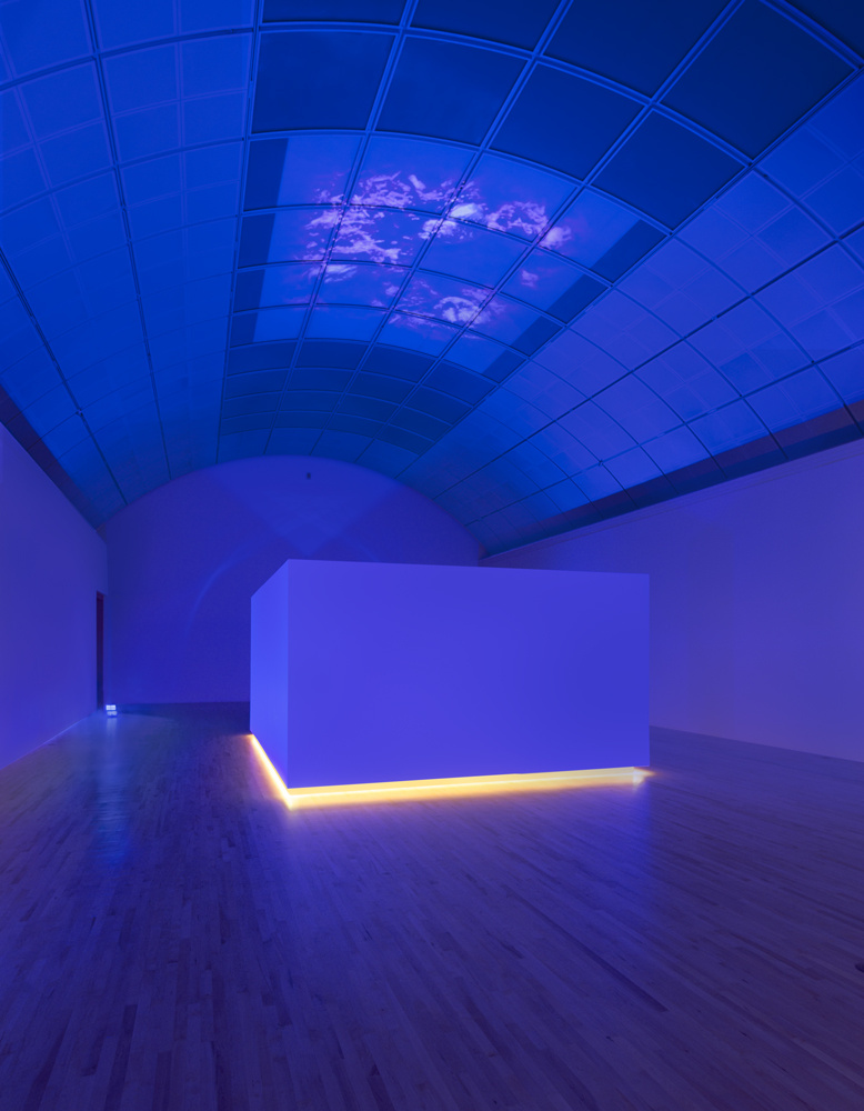A photograph of a cobalt blue room with a domed panel ceiling. Clouds appear in the middle of the dome. There is a large white cube that has light emanating from the bottom of it. Cobalt blue from the walls reflects onto the cube.