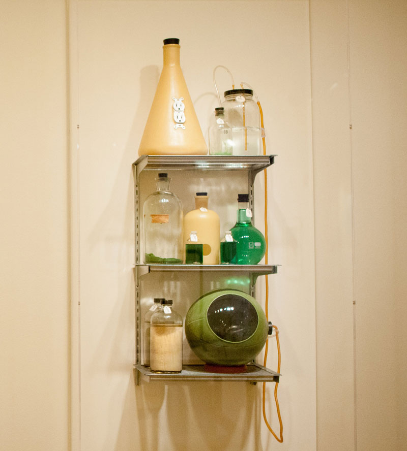 A metal shelf with various scientific beakers. The top shelf holds a bottle with an outlet and tubes coming out of it. The middle shelf contains jars of different shapes with green liquid. The bottom shelf has a sphere with a tube connecting another jar.