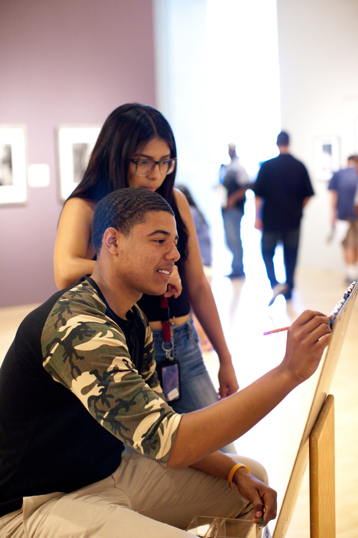 A seated college student in tan pants and black shirt with camouflage detail paints on a sawhorse, whist smiling. Another college student peers over his shoulder observing. Figures walk in the background of the gallery space.