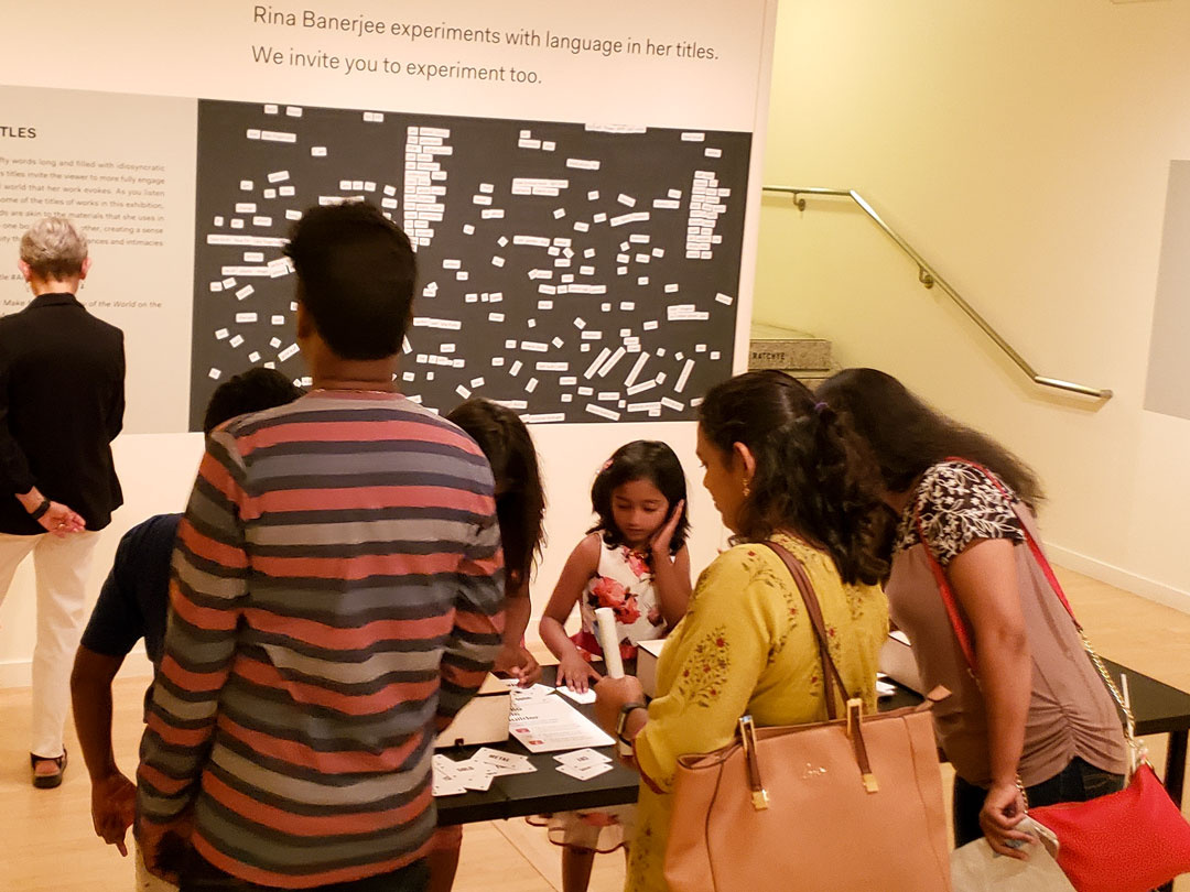 A family with three children is gathered around a table deeply engaged in a hands-on activity. A visitor behind them reads a text panel explaining a magnetic poetry activity.
