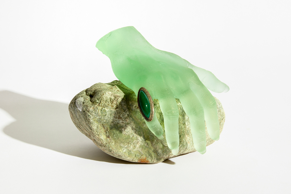 A light green translucent hand wears a green stone ring. It sits on a large stone against a white background.