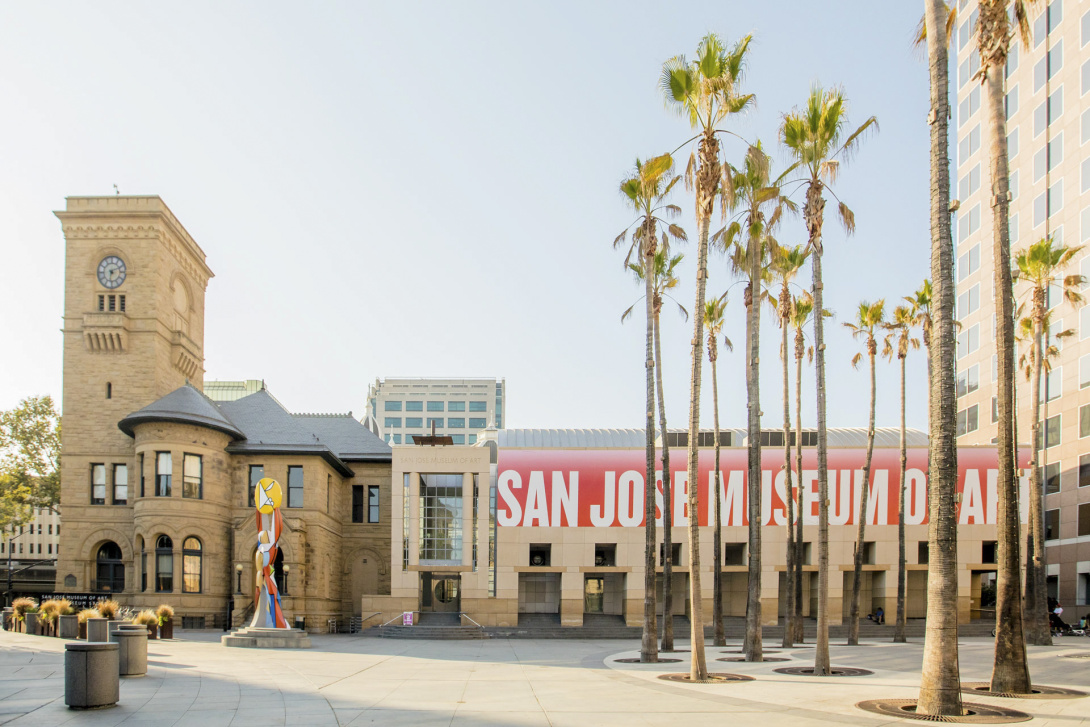  The image shows the exterior of the San Jose Museum of Art. The building has two distinct sections: a historic section on the left with a clock tower and stone architecture, and a modern section on the right with a large banner that reads "SAN JOSE MUSEUM OF ART." Palm trees line the front of the museum, and a colorful abstract sculpture is visible in the foreground. The scene is set in a sunny, urban environment.