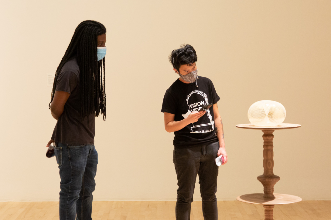 Two visitors looking at an abstract glass work on a wooden pedestal.