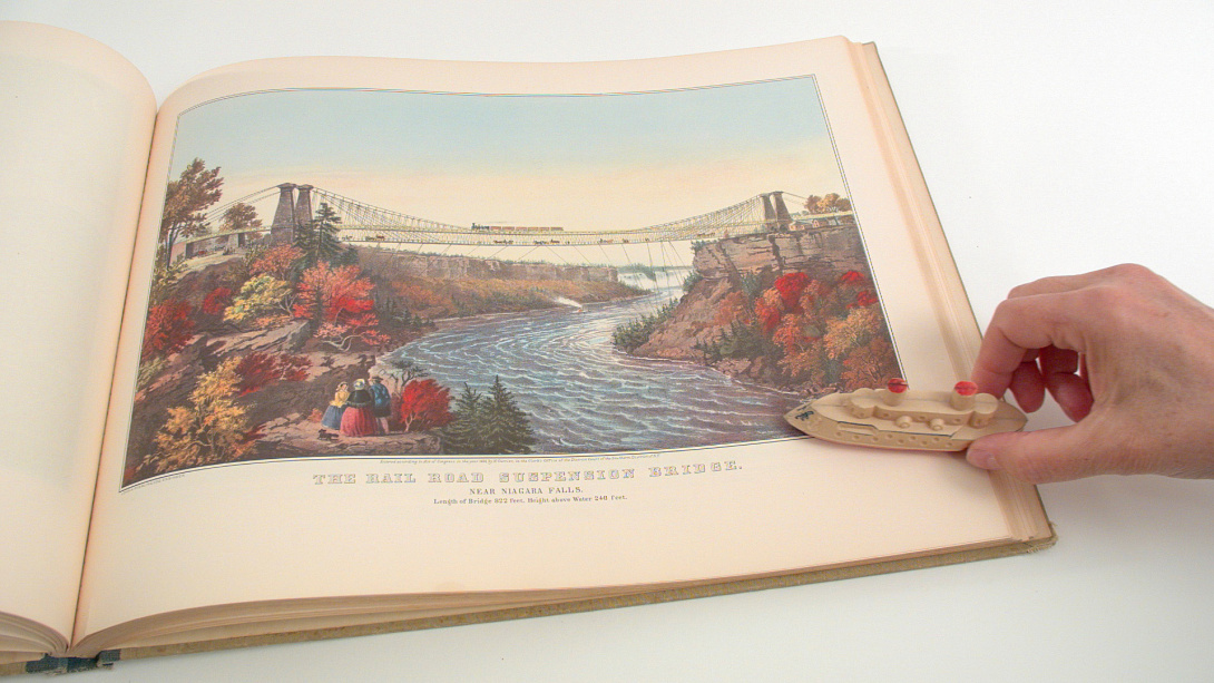 A photograph of a hand guiding a toy boat across an illustration of a river with mountains on either side. The illustration is somewhat mauve tinted and on the right hadn't side of a large hardcover book.