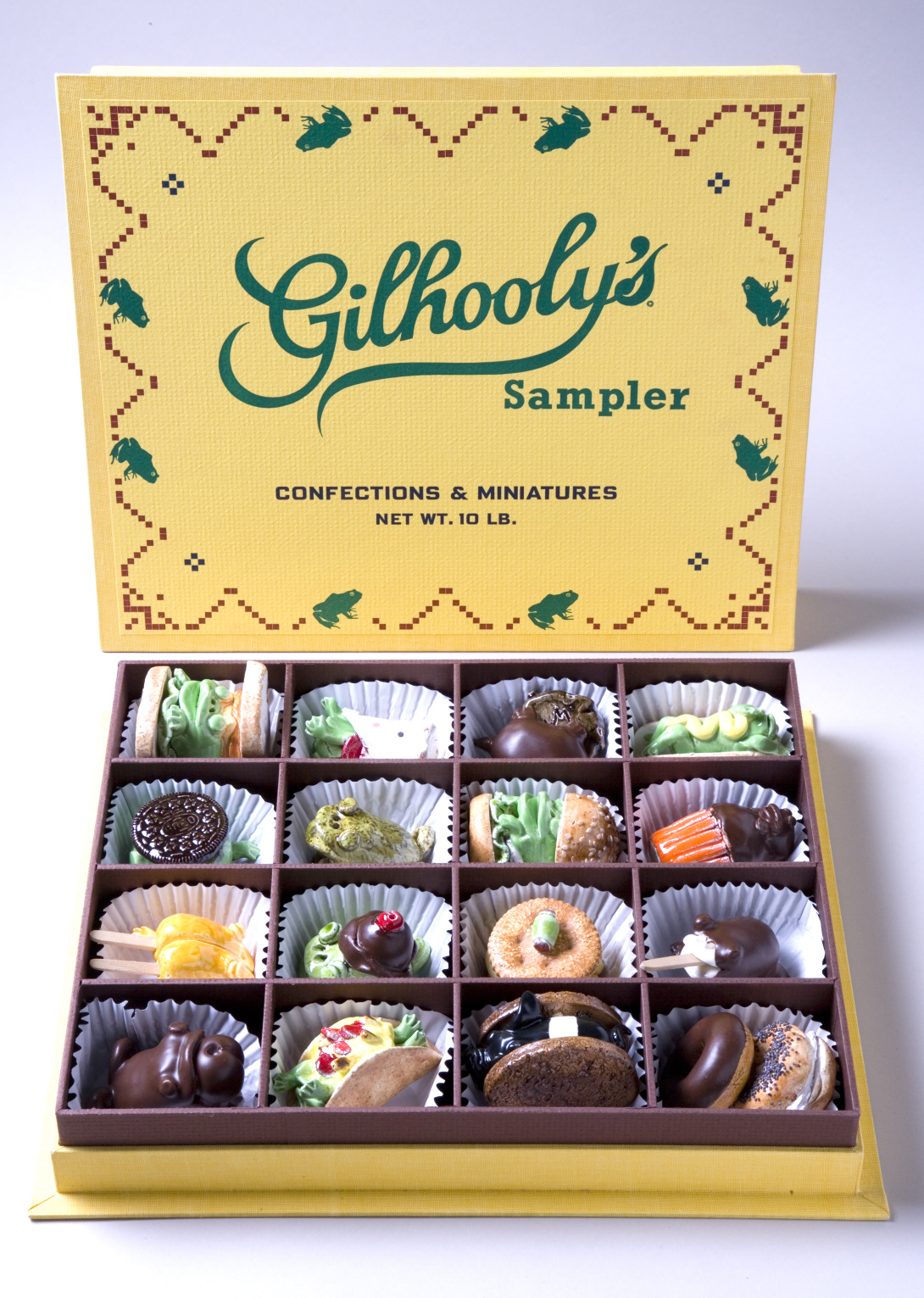 A ceramic yellow candy sampler box that reads "Gilhooly's" in green on the interior of the open lid. Inside the box are a number of smaller ceramics that look like chocolate truffles.