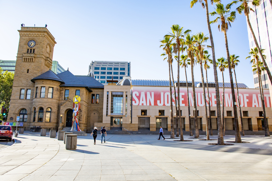 The exterior of a long building that has a large red banner that reads “San Jose Museum of Art.” A circle of palm trees are to the left of the building, with the historic clocktower visible to the far end of the museum. 