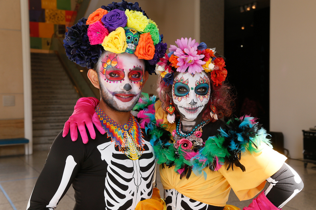 A man and woman stand facing the camera. They are dressed for dia de los muertos, wearing la Calavera, or sugar skull face paint. They each have flowers on top of their head and they are both wearing black tops with white skeleton outlines.