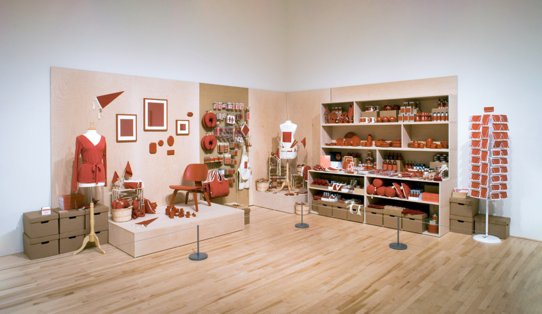 An installation that appears to be multiple shopping experiences in one. Everything is the same uniform rusty brown: college dorm furniture is on a riser; shelves filled with housewares; postcard rack; crafting supplies; and more.