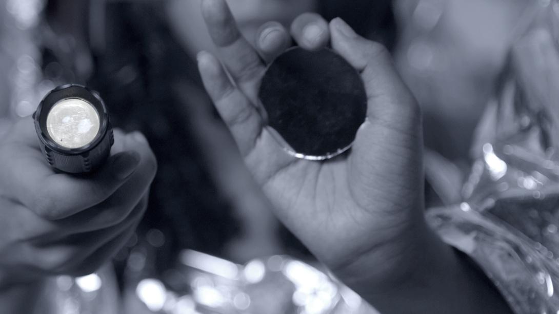 two hands with one holding a flashlight pointing towards the viewer and one holding small circular mirror on the palm