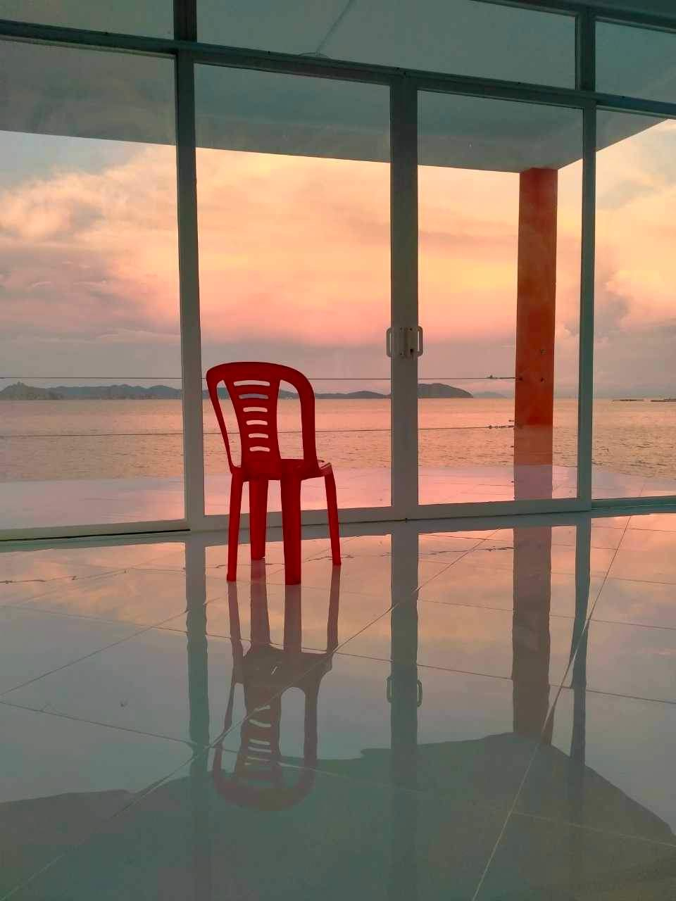 A red plastic chair sits in front of a large glass window with a view of the ocean and distant islands at sunset. The tiled floor reflects the chair and the soft hues of the sky.