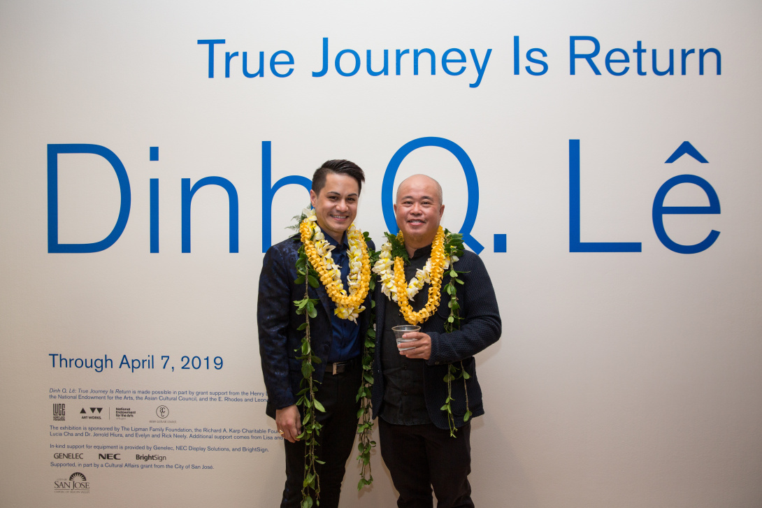 Two men wearing leis stand in front of a wall with large blue text that reads "True Journey Is Return" and "Dinh Q. Lê." The exhibition details and sponsors are listed below. One man is holding a drink, and both are smiling at the camera.